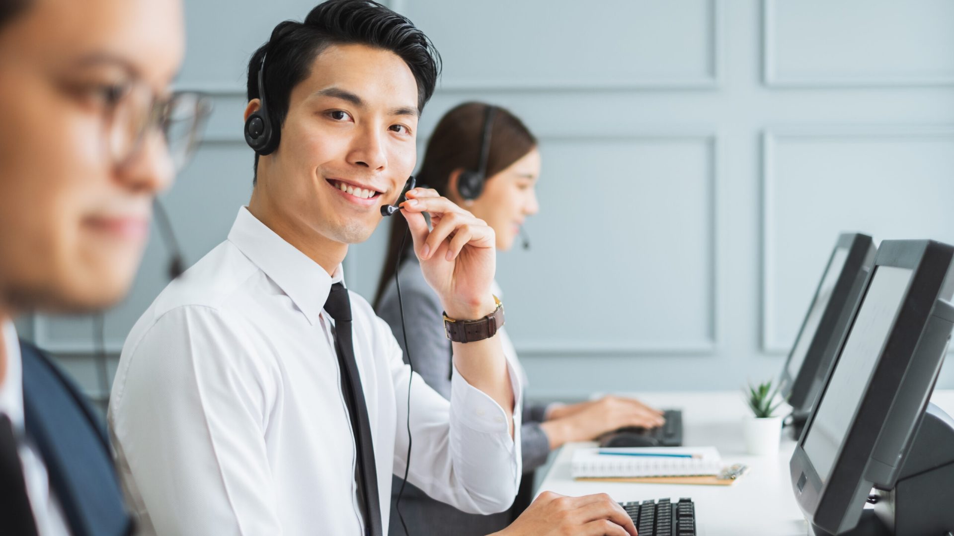 Asian call center team, customer service, telesales in formal suit wearing headset or headphone talking with customer in modern office